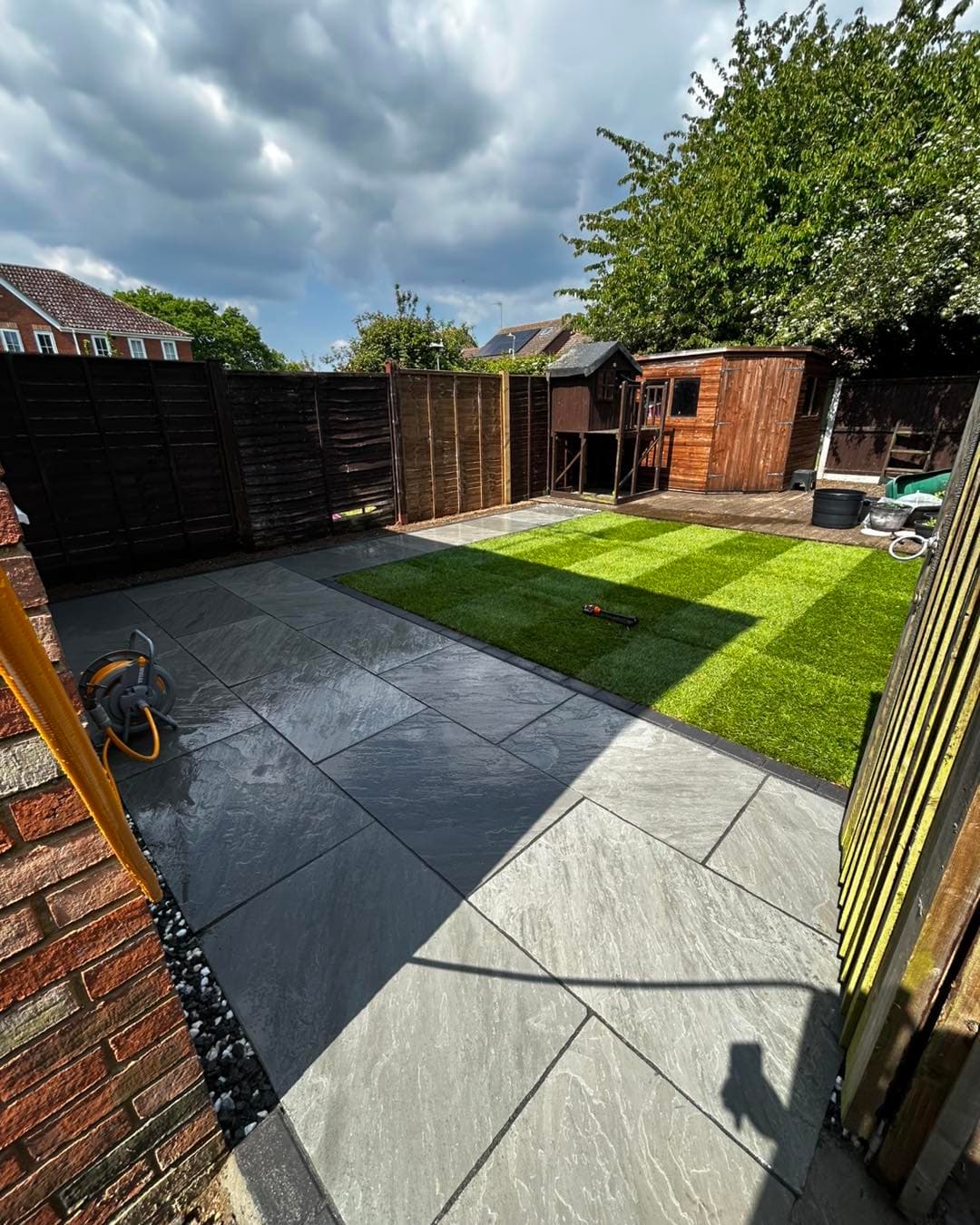Grey stone patio and vibrant green lawn in a fenced backyard with a wooden shed.