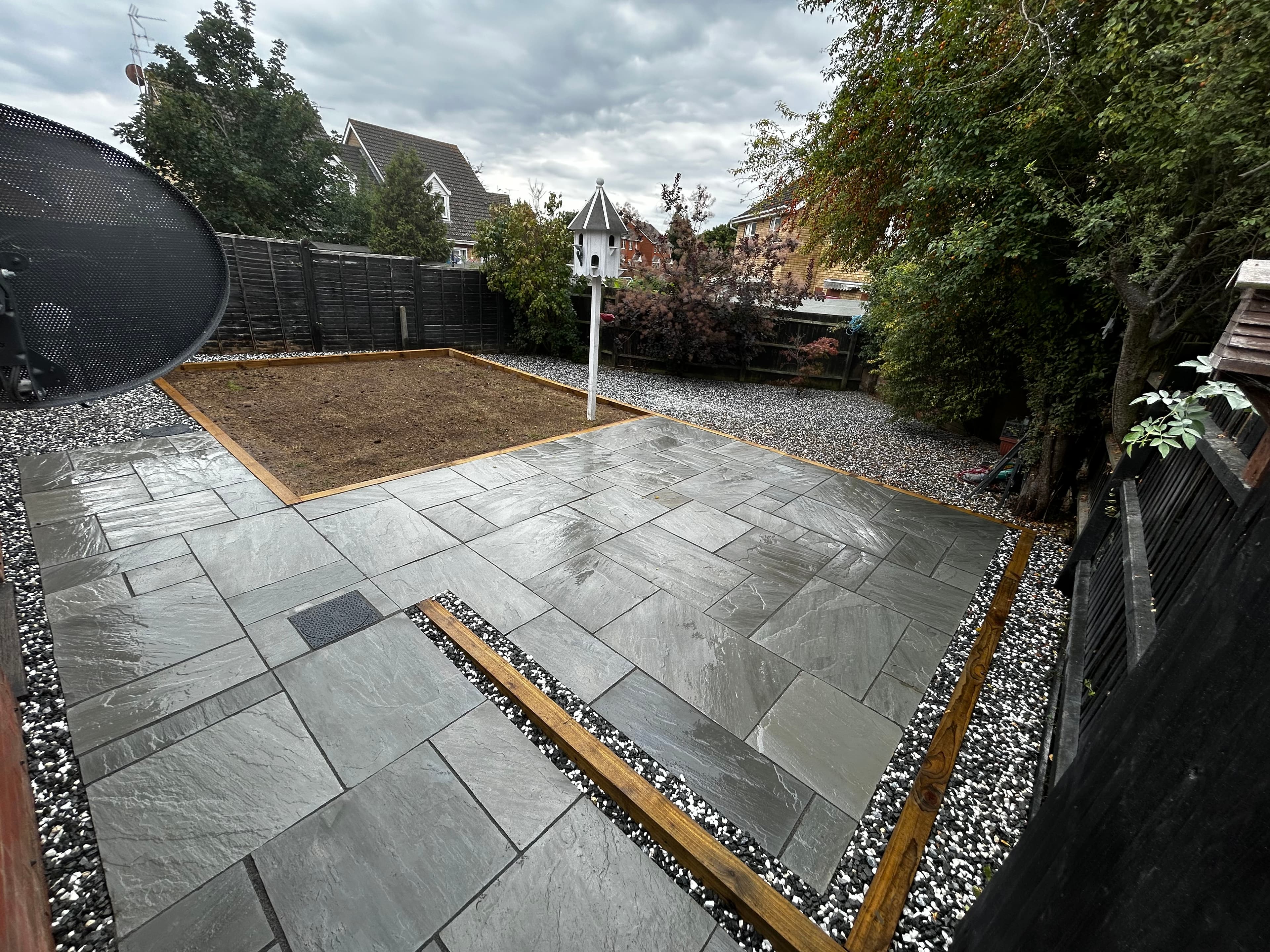 Modern grey stone patio and bare soil garden bed surrounded by gravel in a backyard.
