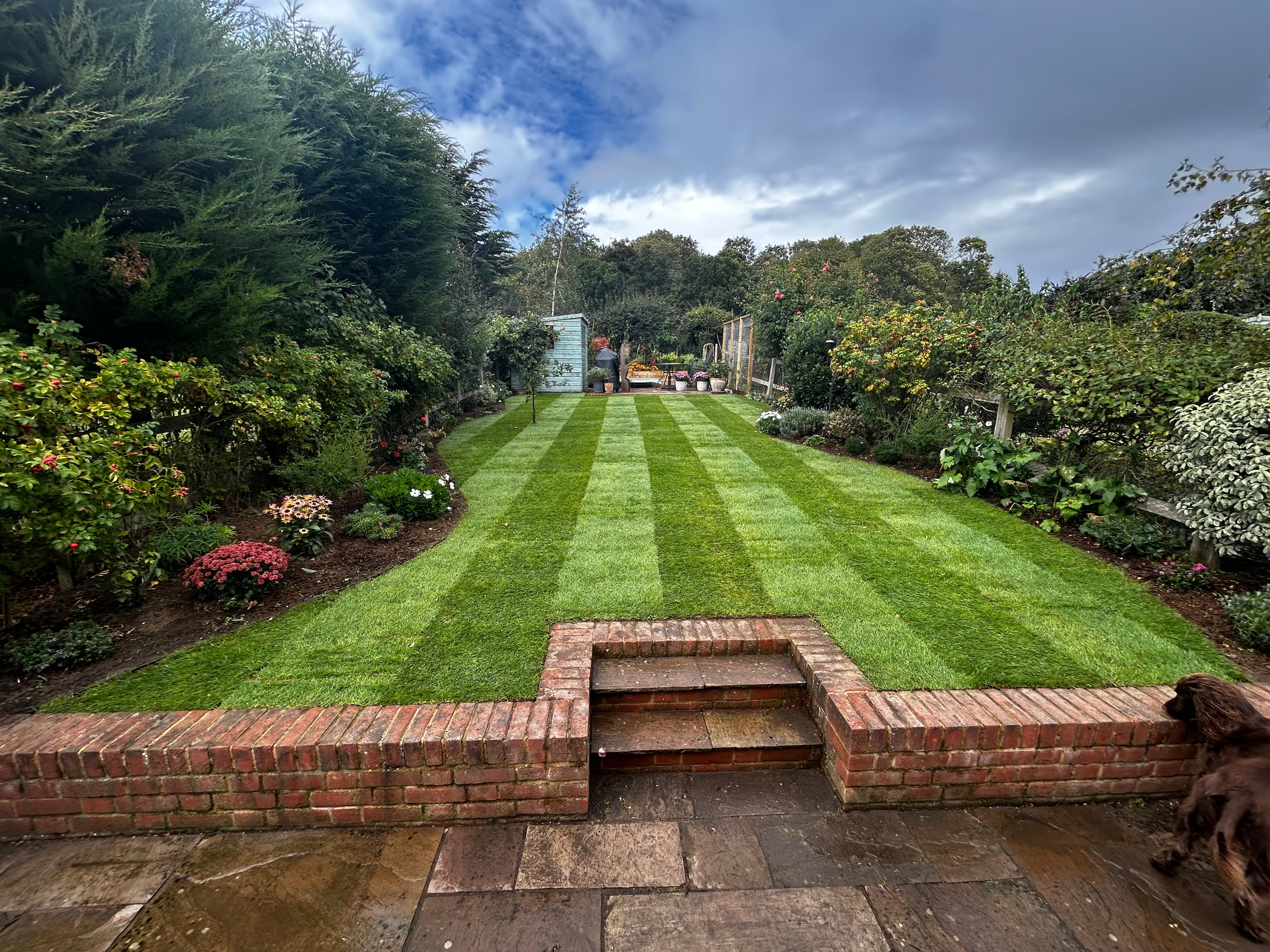 Striped green lawn bordered by lush gardens, viewed from a stone patio with brick steps.