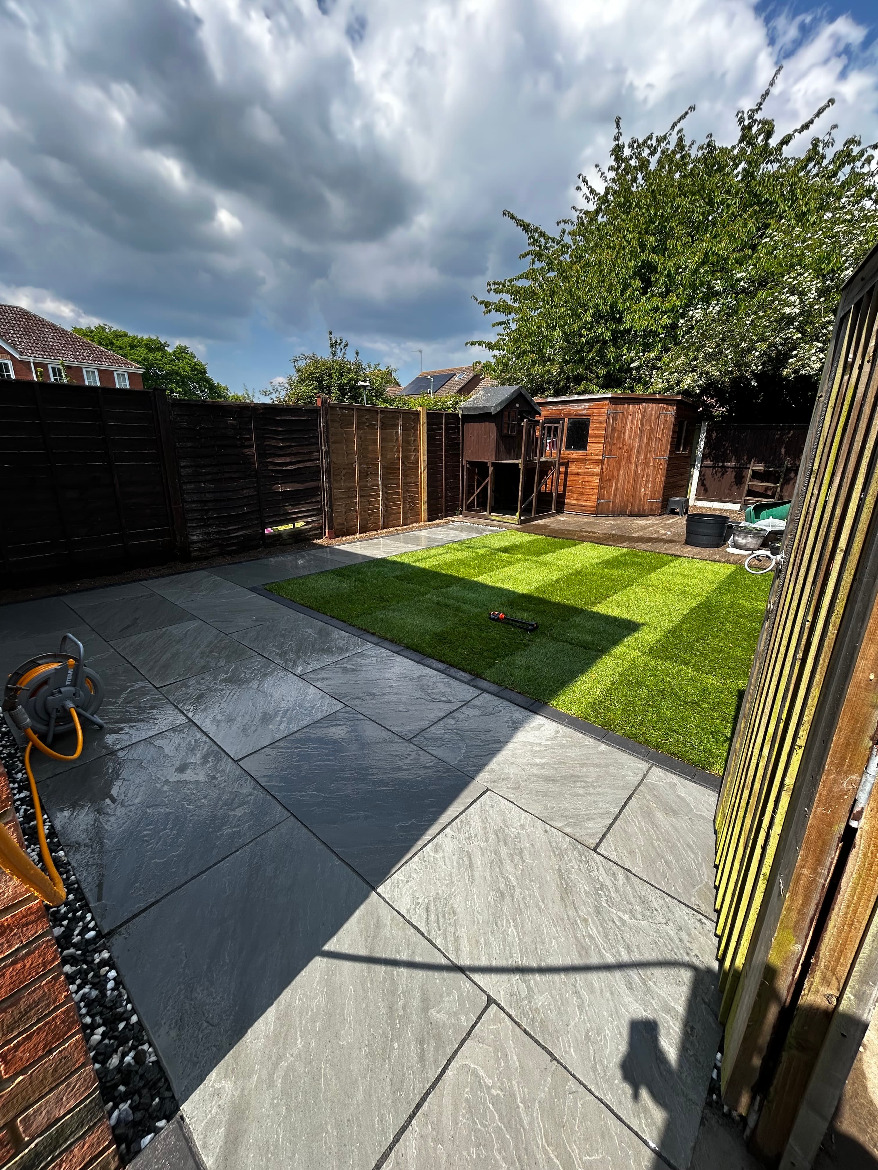 Modern backyard with grey stone patio, striped artificial lawn, wooden shed, and large tree.