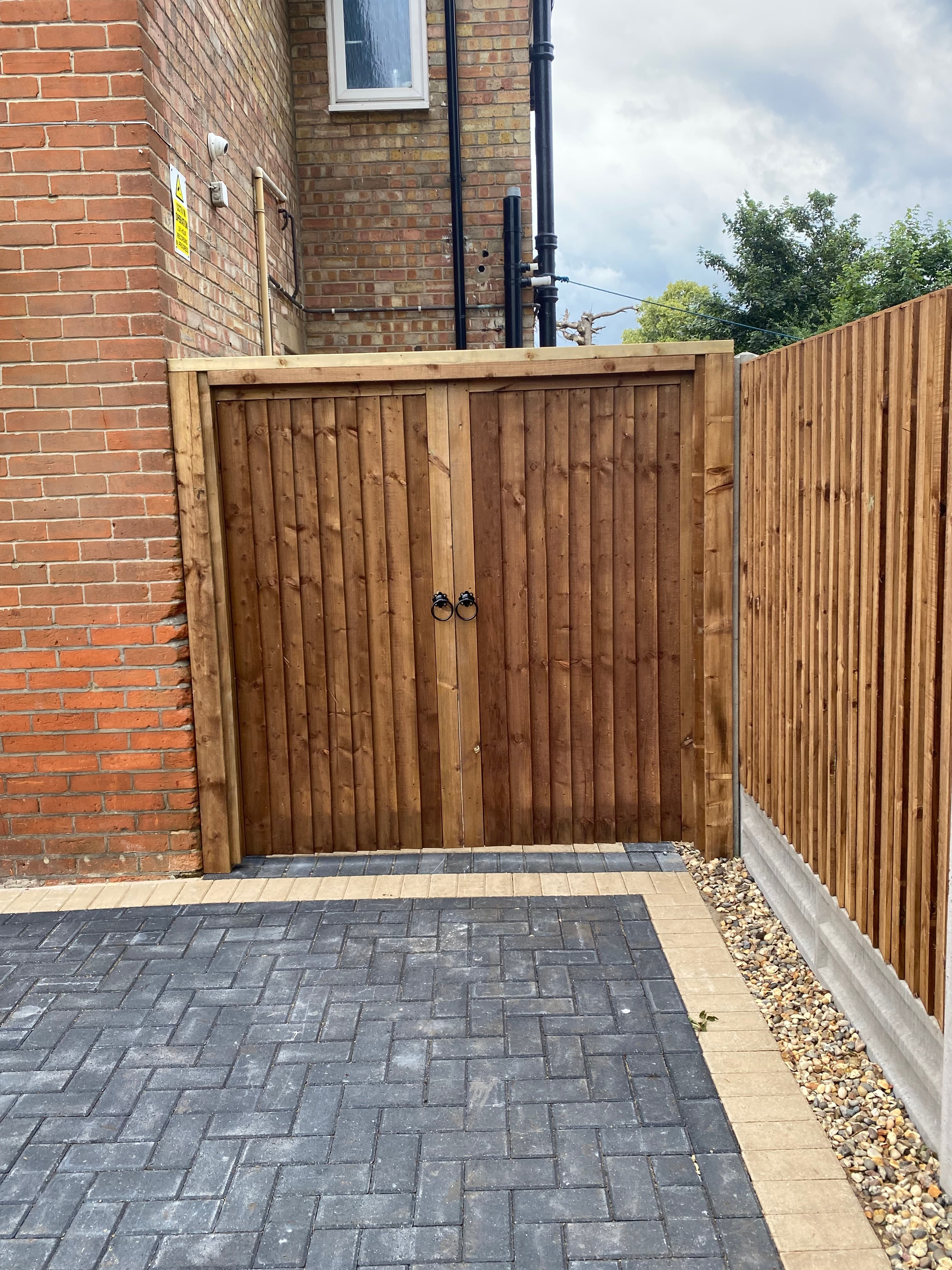 Wooden storage shed with double doors against a brick wall next to a paved driveway.