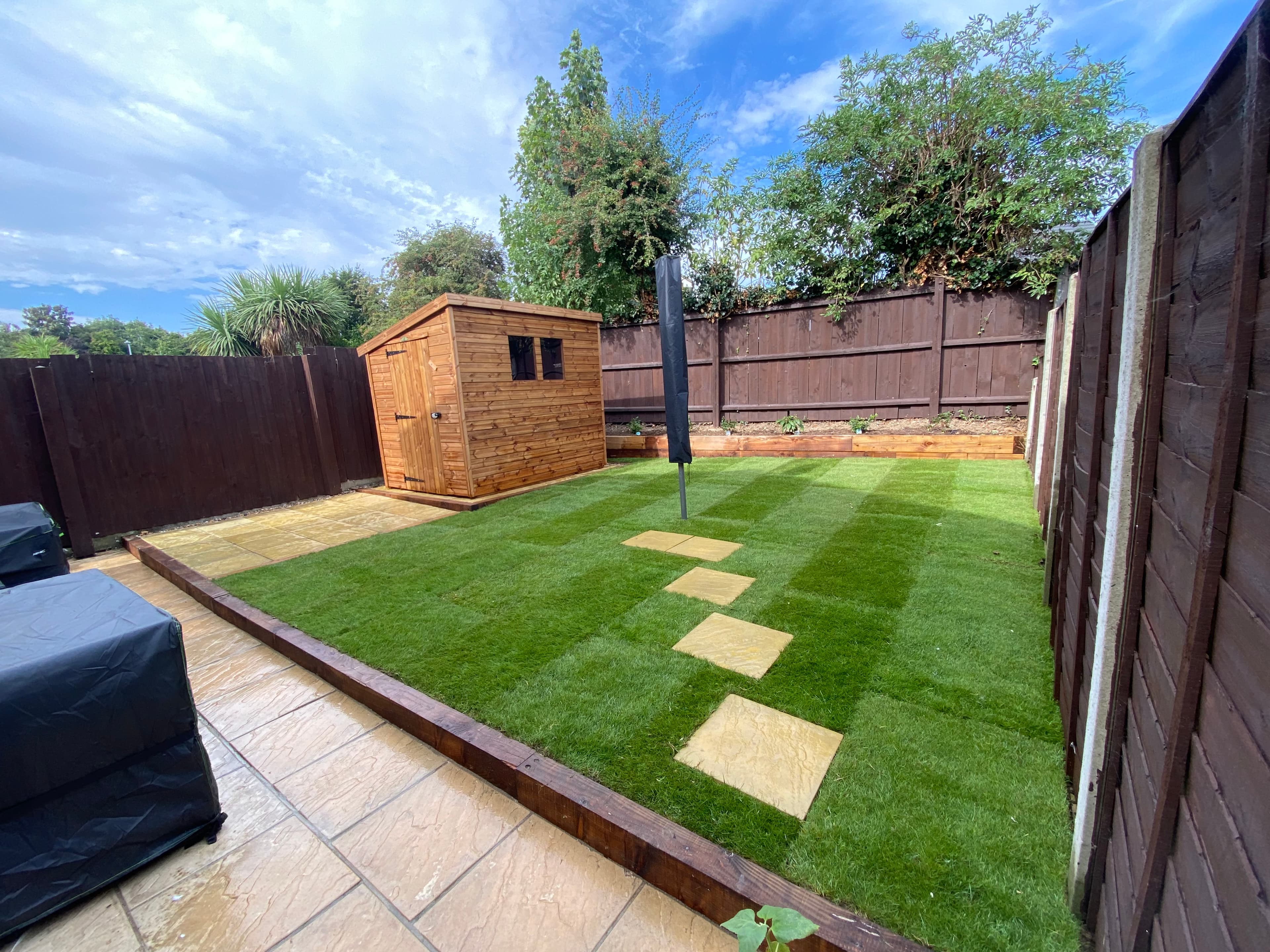 Landscaped backyard with a wooden shed, striped green lawn, stepping stones, and stone patio.